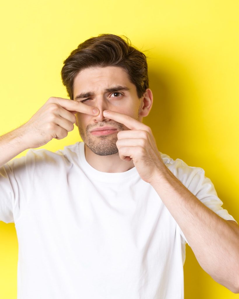 Young man squeezing pimple on nose, standing over yellow background. Concept of skin care and acne.